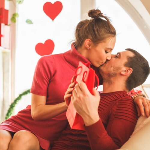 A woman in red and a man in red. The woman is sitting on his lap kissing him. Red Hearts Are In The background.